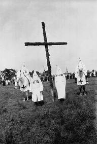An old black and white picture showing four KKK members gathered in their traditional head dress and robes under a wooden cross, two of the members are on horse back 