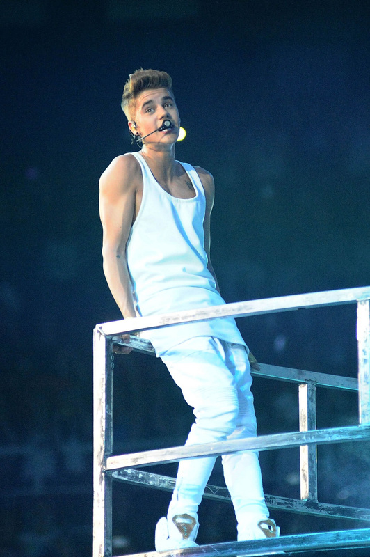 Picture of Justin Bieber in a white vest, jeans and high-tops, leaning on some railings whilst in concert.